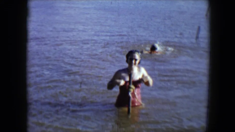 A 1959 home-movie frame shows the woman waist-deep in muddy water, centered near a pole with ripples and a swimmer behind her.
