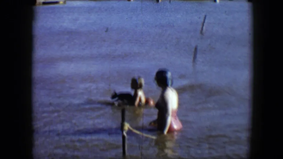 1959 Disneyland frame showing a woman in a red swimsuit near another swimmer, a small dog, posts, and rope in muddy water.