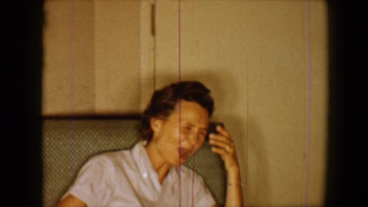 1959 8mm home-movie frame shows an adult woman in a light blouse seated indoors, smiling with one hand raised near her face.