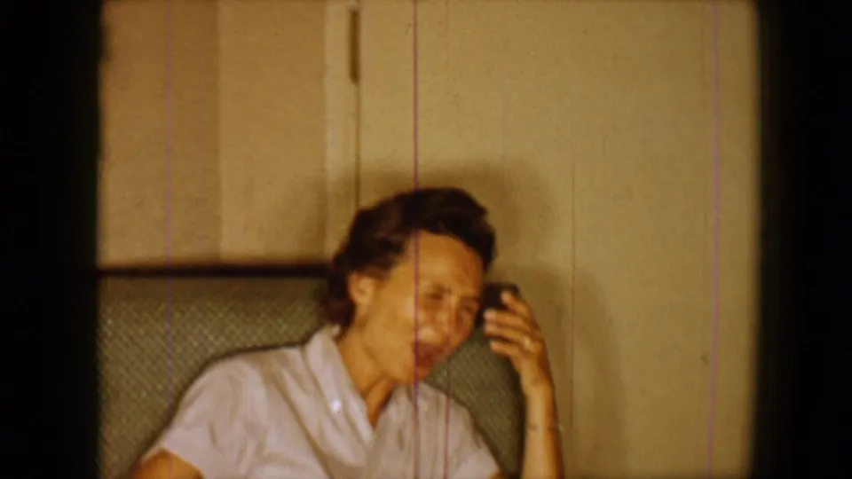1959 8mm home-movie frame shows an adult woman in a light blouse seated indoors, smiling with one hand raised near her face.