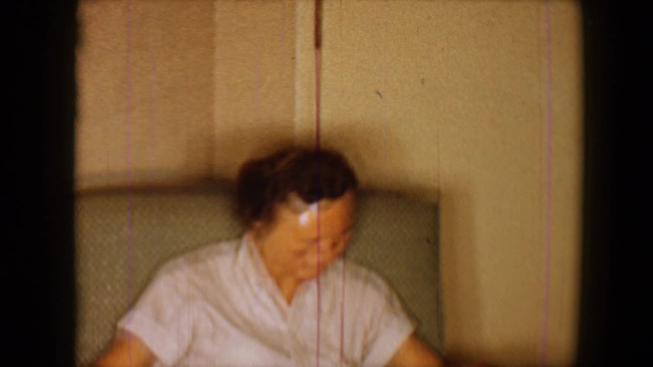 Adult woman in a light blouse seated indoors, looking down beside a chair back.