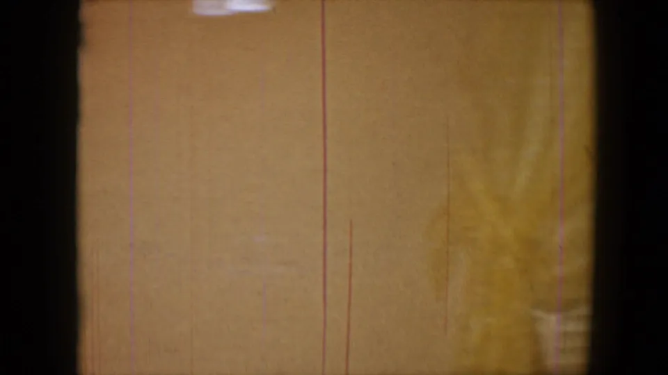 A transitional 8mm frame shows a pale interior wall or surface with visible film scratches, grain, and partial room detail.