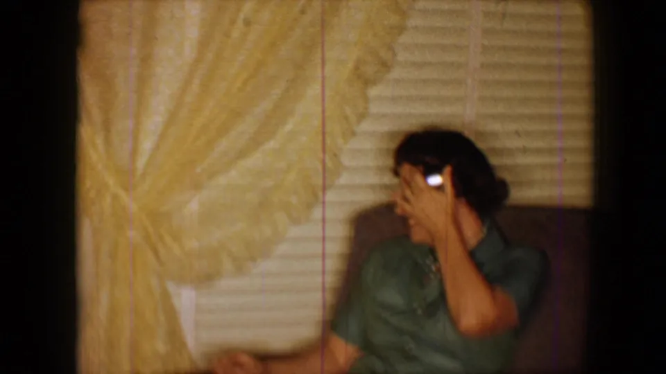 1959 8mm home-movie frame shows an adult woman in a green top seated near blinds, covering part of her face while laughing or reacting.