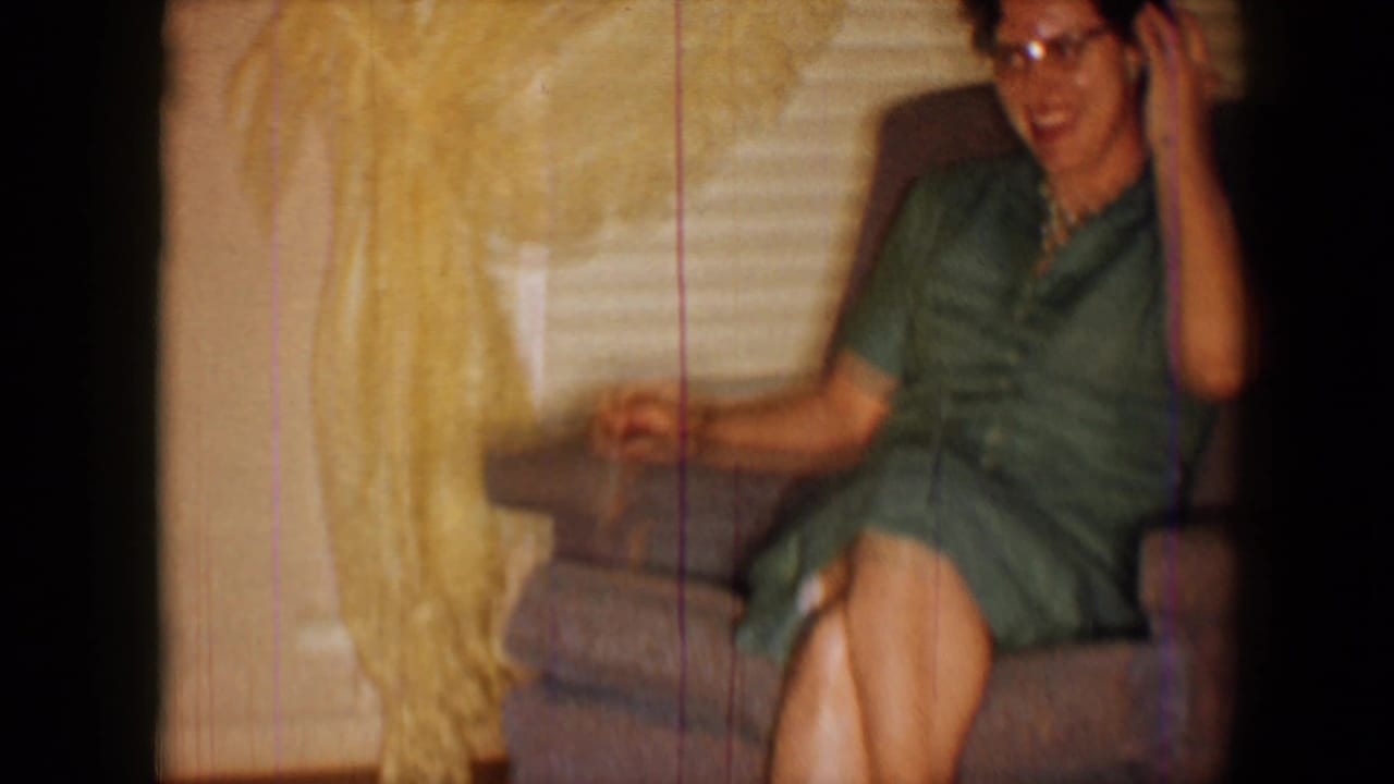 1959 8mm home-movie frame of a woman in a green dress seated in a brown recliner in a sunlit private living room.