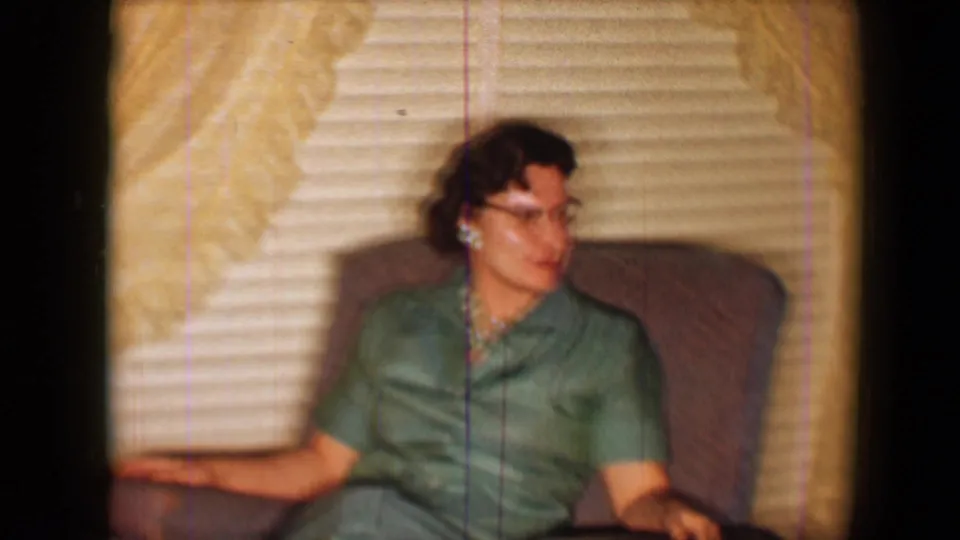 1959 8mm frame of a woman in a green dress looking off camera while seated in a brown recliner in a living room.