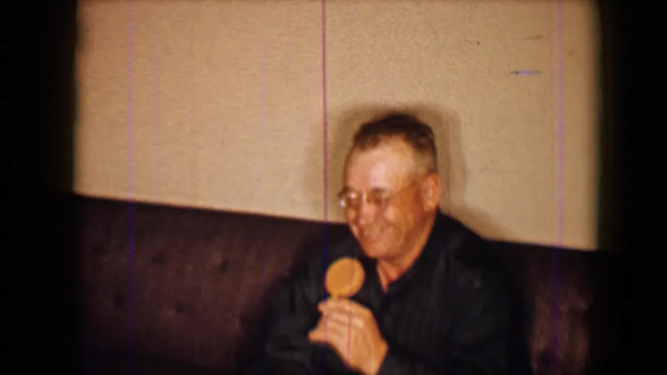 1959 home-movie frame showing an older man in a dark shirt seated against a plain wall, holding a small round object.