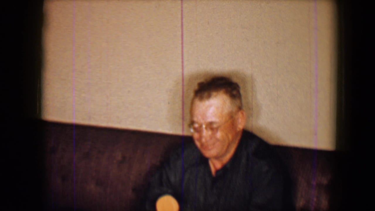 The man remains seated on the couch at the 1959 Disneyland setting, holding the round lolly near his lap in soft 8mm focus.