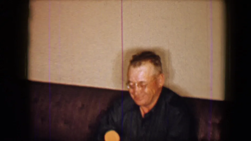 The man remains seated on the couch at the 1959 Disneyland setting, holding the round lolly near his lap in soft 8mm focus.