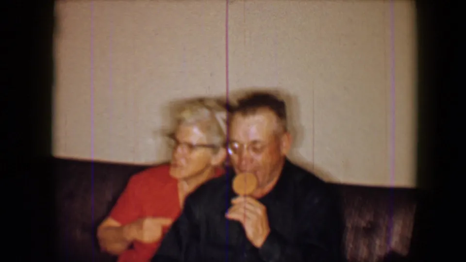 The woman watches and gestures while the man raises the round lolly to his mouth on the couch in the 1959 Disneyland footage.