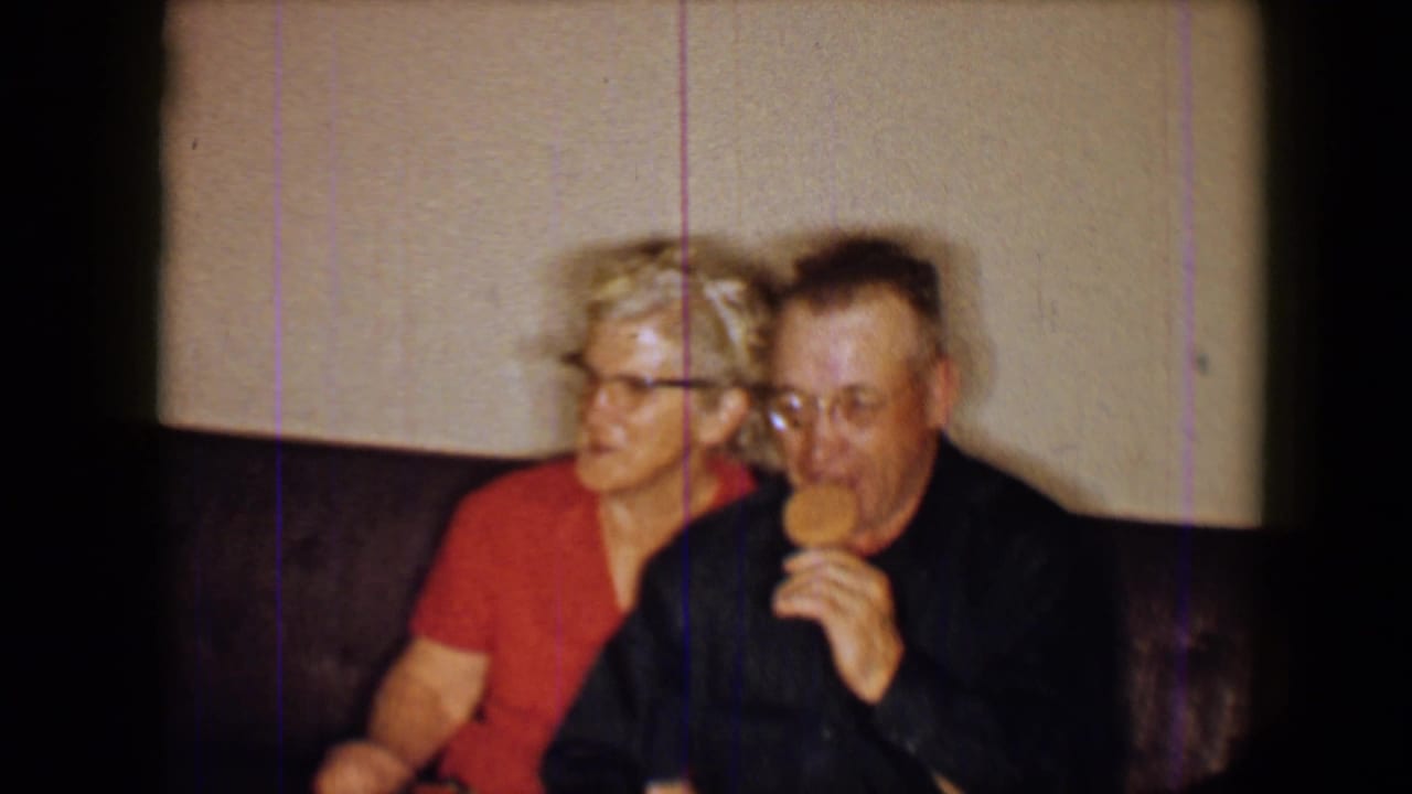 Older man eating a lollipop beside a woman in red on a sofa.