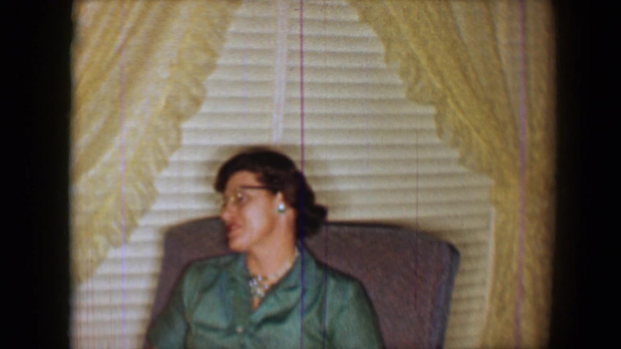 1959 home-movie frame of a woman in a green blouse and glasses seated indoors before curtains and venetian blinds.