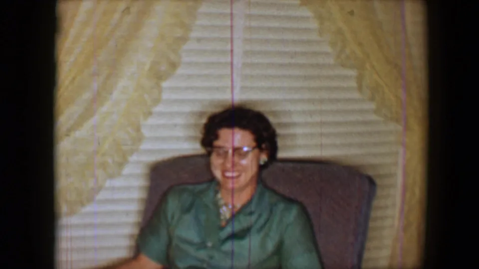 1959 8mm frame of a woman smiling in a green blouse while seated in a patterned armchair indoors.