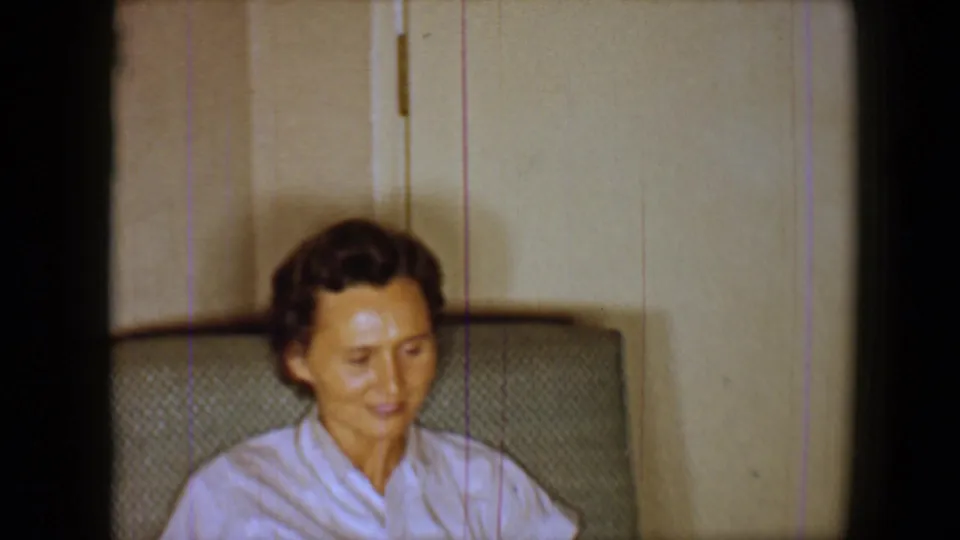1959 home-movie frame of a woman in a pale top seated indoors, looking downward near a paneled wall.