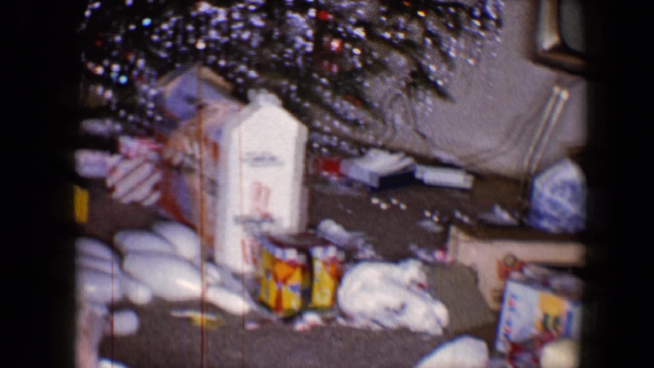1959 8mm home-movie frame of a holiday living room with tinsel, opened gifts, packaging and a tabletop television.