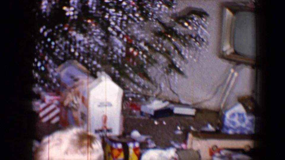 1959 home-movie frame with tinsel branches overhead, opened Christmas packaging on the floor and a small television nearby.