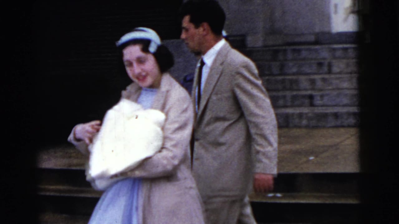 Smiling woman carries a bundled baby beside a man in a beige suit near steps.