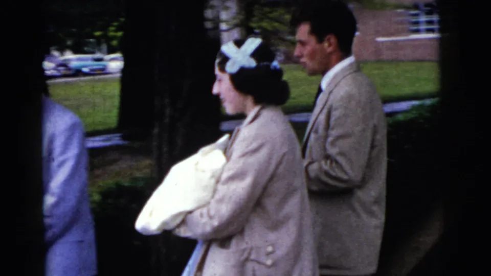Side view of the woman carrying a swaddled newborn outdoors, with a man in a beige suit following behind in the 1961 Gary footage.