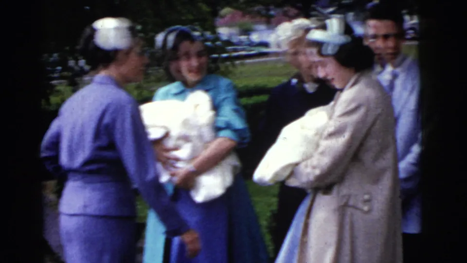 Adults gather outdoors in Gary, Indiana, 1961, with two swaddled newborns held among women and a man near a lawn and parked cars.