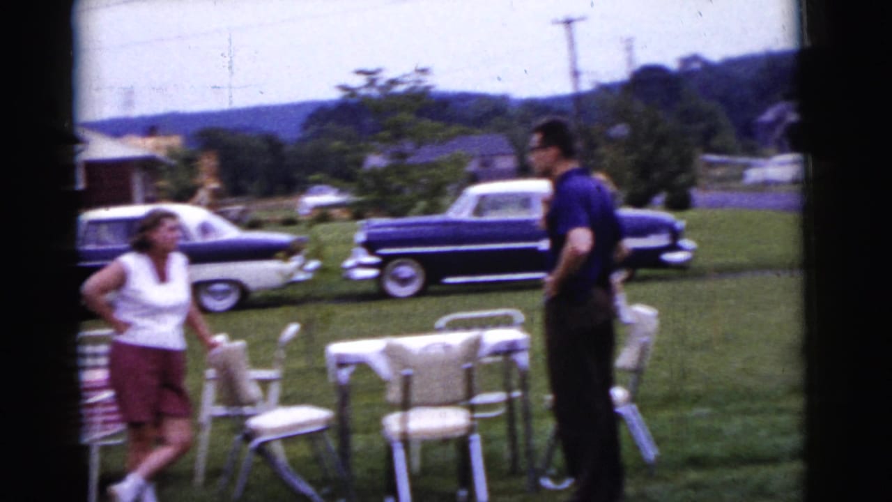 Woman and man standing in a yard near empty tables and folding chairs with parked cars behind them.