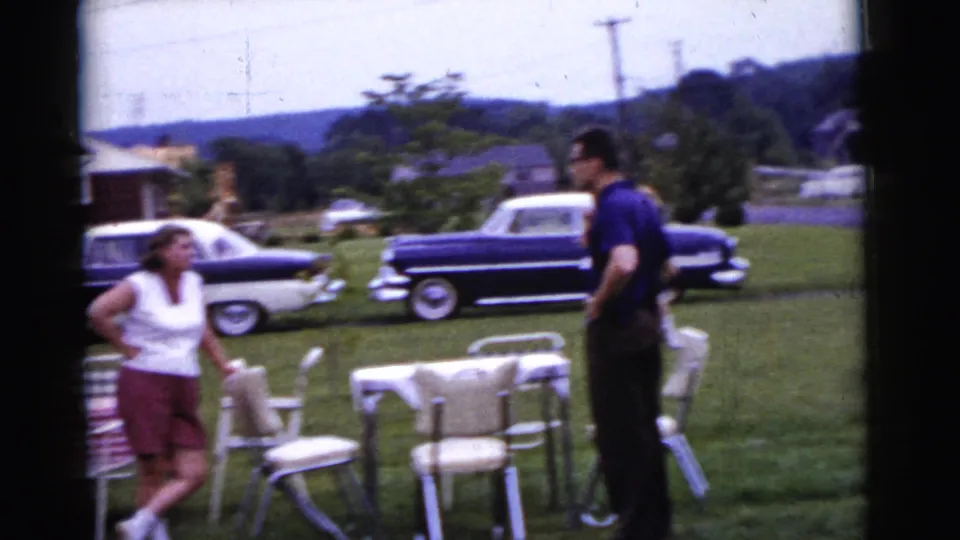 1961 Gary, Indiana home-movie frame of two adults in a suburban yard with empty tables, chairs, lawn, house, and period cars.