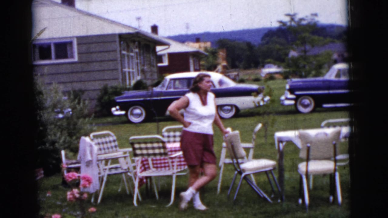 Woman standing near folding chairs and tables in a lawn with two-tone cars and a house in the background.