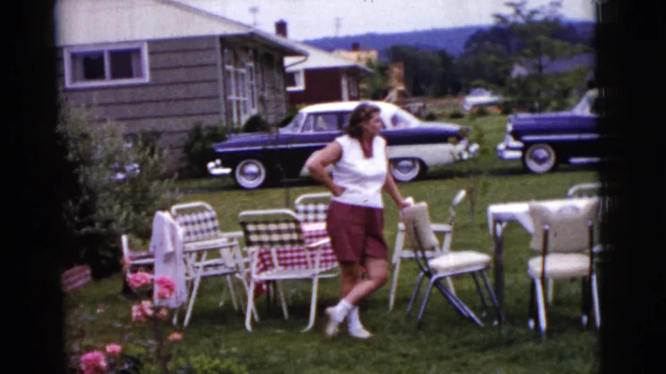 1961 Gary, Indiana home-movie still of a woman near arranged backyard seating, flowers, a house, and parked mid-century cars.