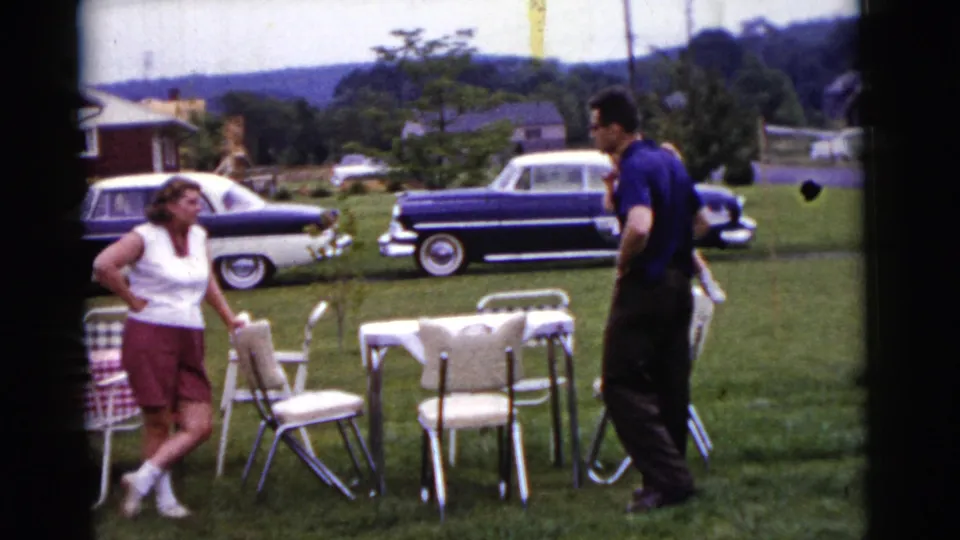 1961 Gary, Indiana frame with two adults facing each other in a backyard gathering setup with chairs, tables, lawn, and cars.