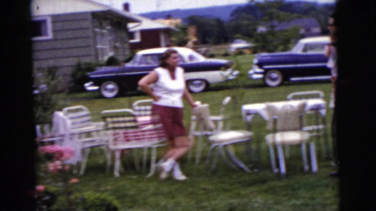Woman standing near white patio furniture in a sunny suburban yard with parked cars behind her.