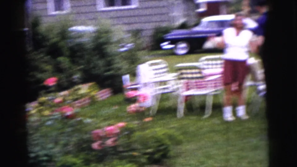 1961 Gary, Indiana frame with handheld blur, showing a woman near white patio chairs, lawn flowers, shrubs, and a dark parked car.