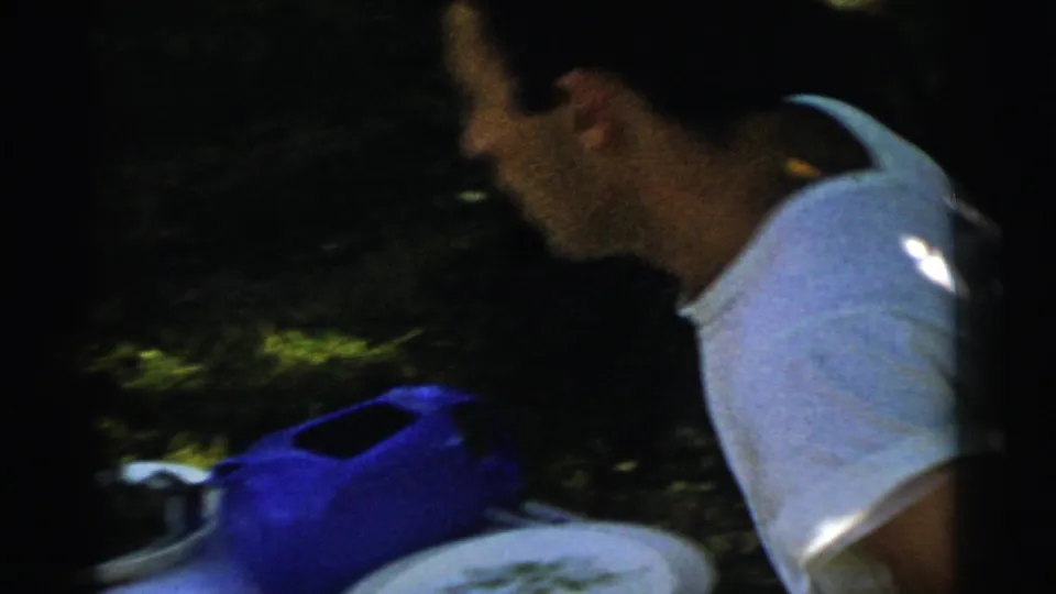 A young man in a white shirt appears in side view near grass and picnic items in Gary, Indiana home-movie footage.