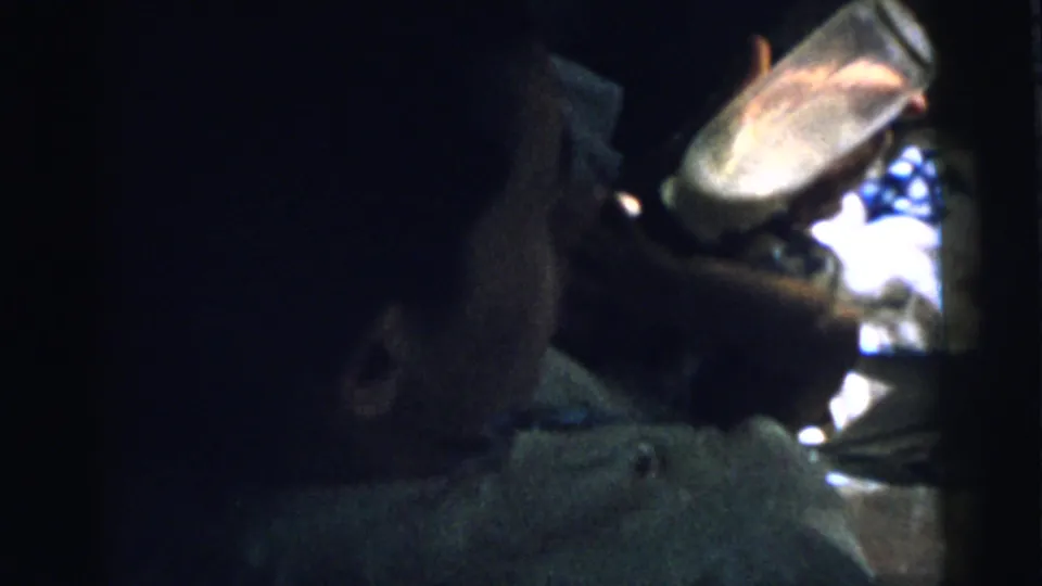 Close 1961 home-movie frame from Gary, Indiana showing bottle feeding, with the infant on bedding and an adult hand in the foreground.