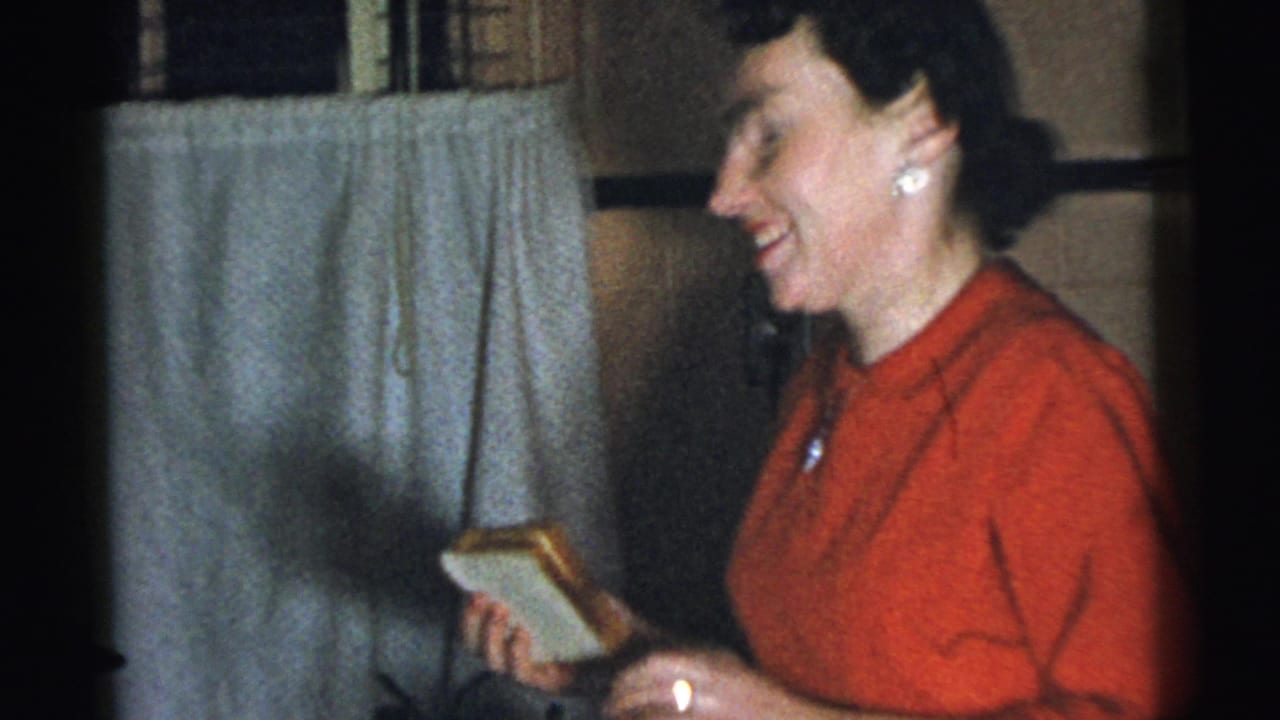 Woman in a red dress laughs while holding a small gift in a dim room.
