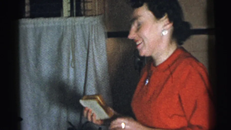 In a 1961 Gary, Indiana home-movie frame, a woman in red laughs while holding a small gift near a curtained window.