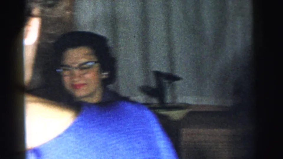 A woman in a blue top and glasses appears near curtains in the same 1961 indoor home-movie setting.