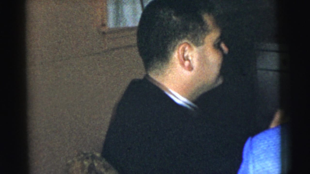 Man in a dark suit stands indoors beside a partially visible person in blue.