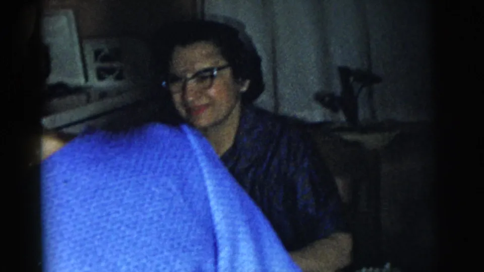 1961 Gary, Indiana home-movie frame showing a seated woman with glasses in a dim room, partly blocked by a person in blue.