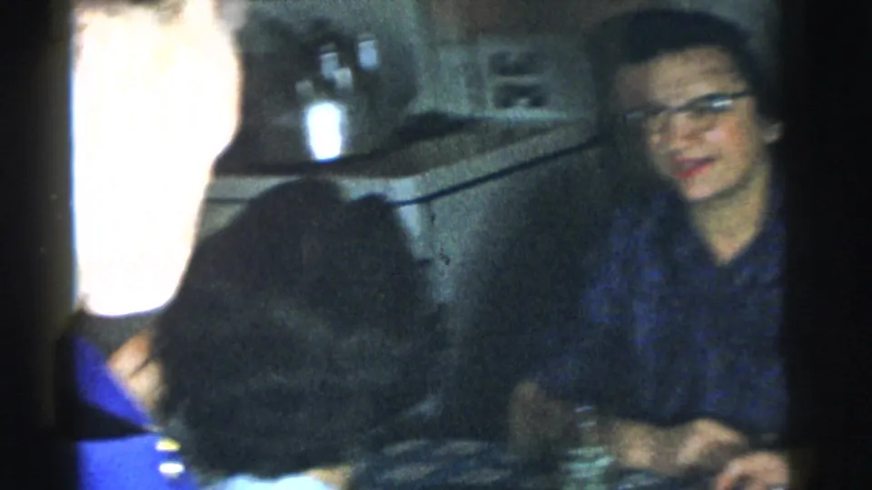 1961 Gary, Indiana frame showing a seated woman with glasses in a private interior, partly obscured by nearby adults.