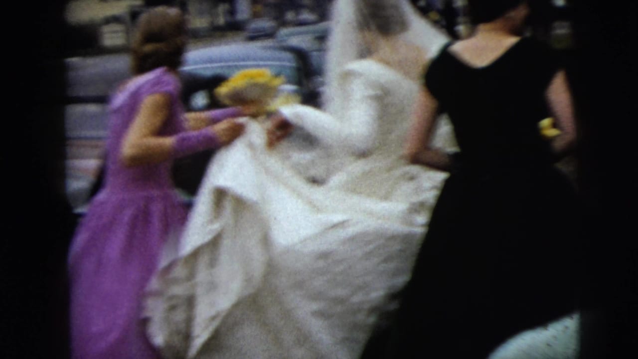 Bride in white gown assisted by bridesmaids on a street.