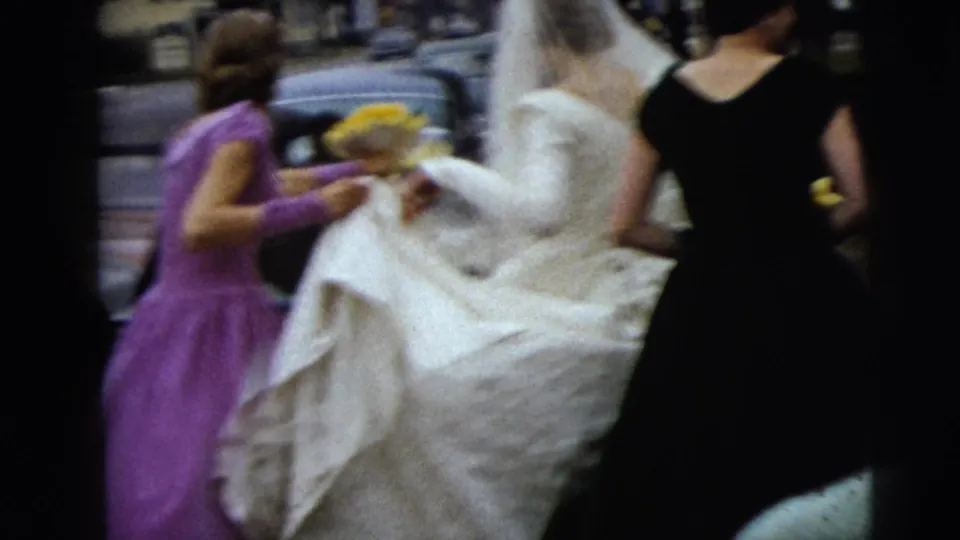 In 1962 Moline, Illinois footage, bridesmaids help a bride in a white gown and veil walk along a street.