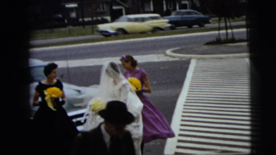 The bride and bridesmaids, one in purple holding yellow flowers, move near a crosswalk with mid-century cars in the background.