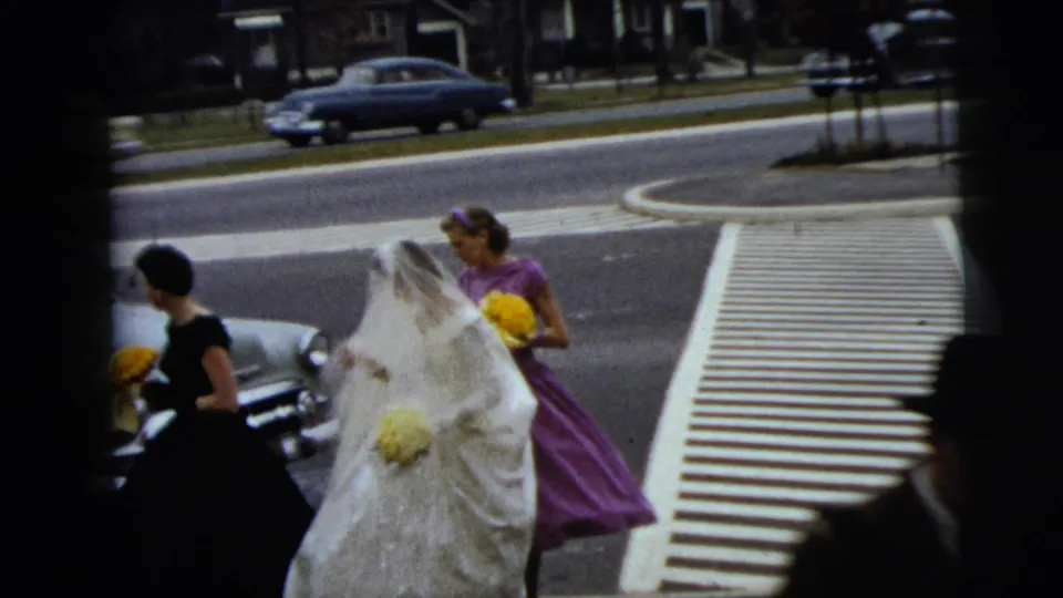In 1962 Moline, Illinois, a bride in a white veil walks with bridesmaids in dark and purple dresses near a marked crosswalk.