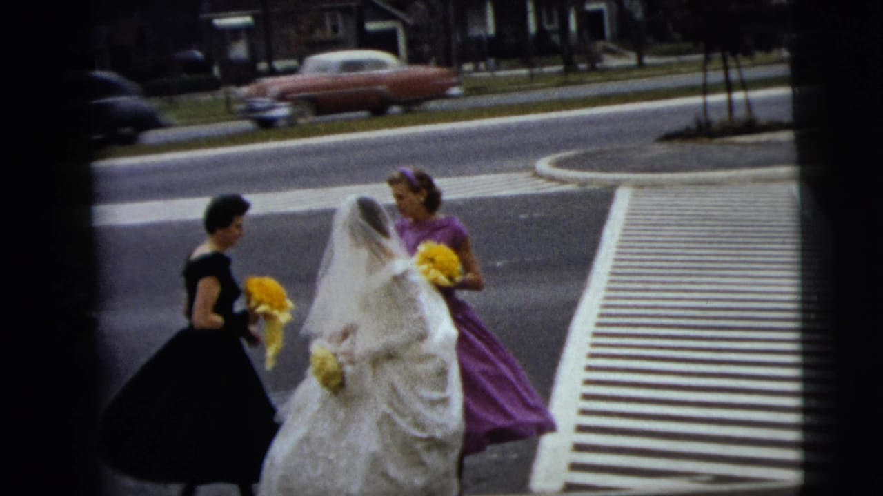 Bride and two bridesmaids stand or walk near the crosswalk while a red car passes in the background.