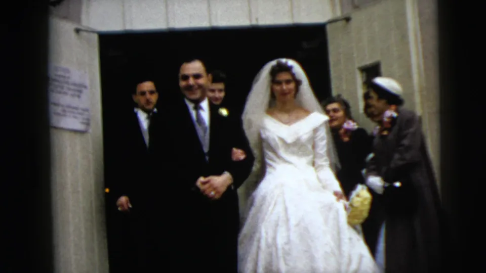A 1962 home-movie frame shows the bride and groom walking from the entrance, with attendants and guests in formal clothing behind them.