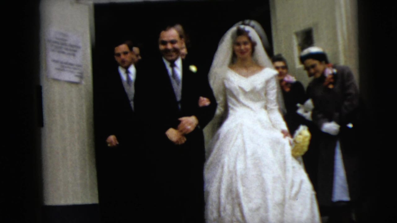In Moline, Illinois, 1962, a bride in a white gown and veil walks arm in arm with the groom down stone church steps as guests follow.