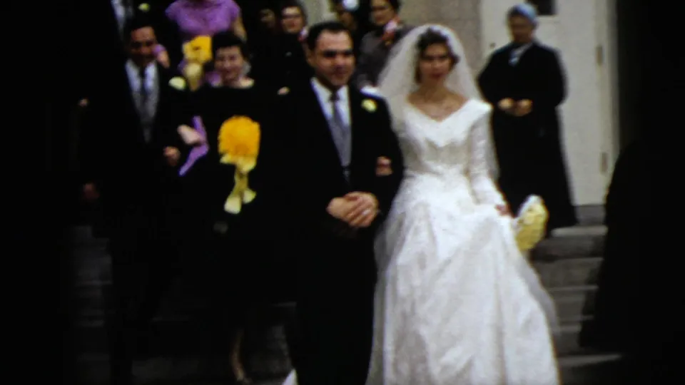 Guests carrying bright flowers gather closely around the bride and groom as the couple moves down the church steps in this 1962 home-movie frame.