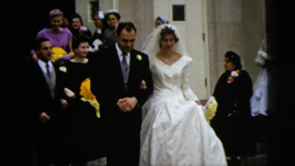 The bride and groom walk forward from the church entrance while family or guests in period formal wear stand nearby, some holding yellow flowers.