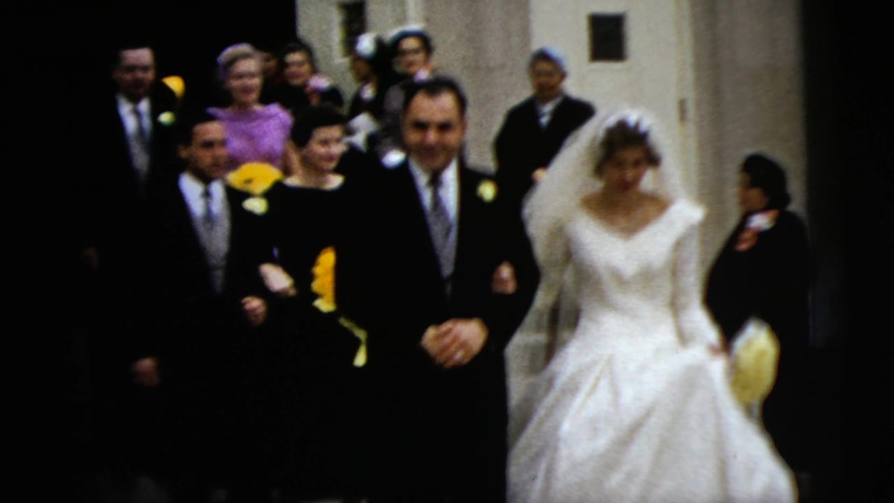 In 1962 Moline, Illinois, a bride in a white gown and veil walks beside a groom in a dark suit as guests follow down church steps.