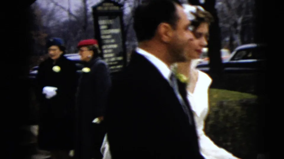 A side view shows the groom and bride walking near the curb, with guests, a signboard, and a parked car in the background.
