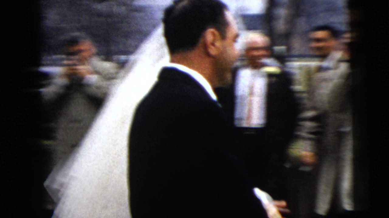 Bride and groom walk past wedding guests toward a street with parked cars.