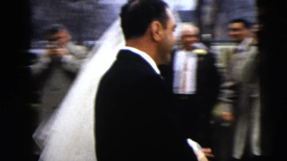 In 1962 Moline, Illinois home-movie footage, a bride in a long veil and groom in a dark suit pass guests on a street.